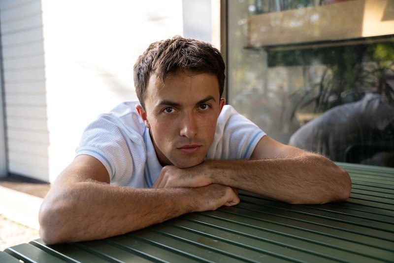 Ein Mann in einem weißen T-Shirt lehnt sich auf einen grünen Tisch und schaut direkt in die Kamera. Im Hintergrund sind unscharfe Pflanzen zu sehen., © Kulturhaus Schwanen