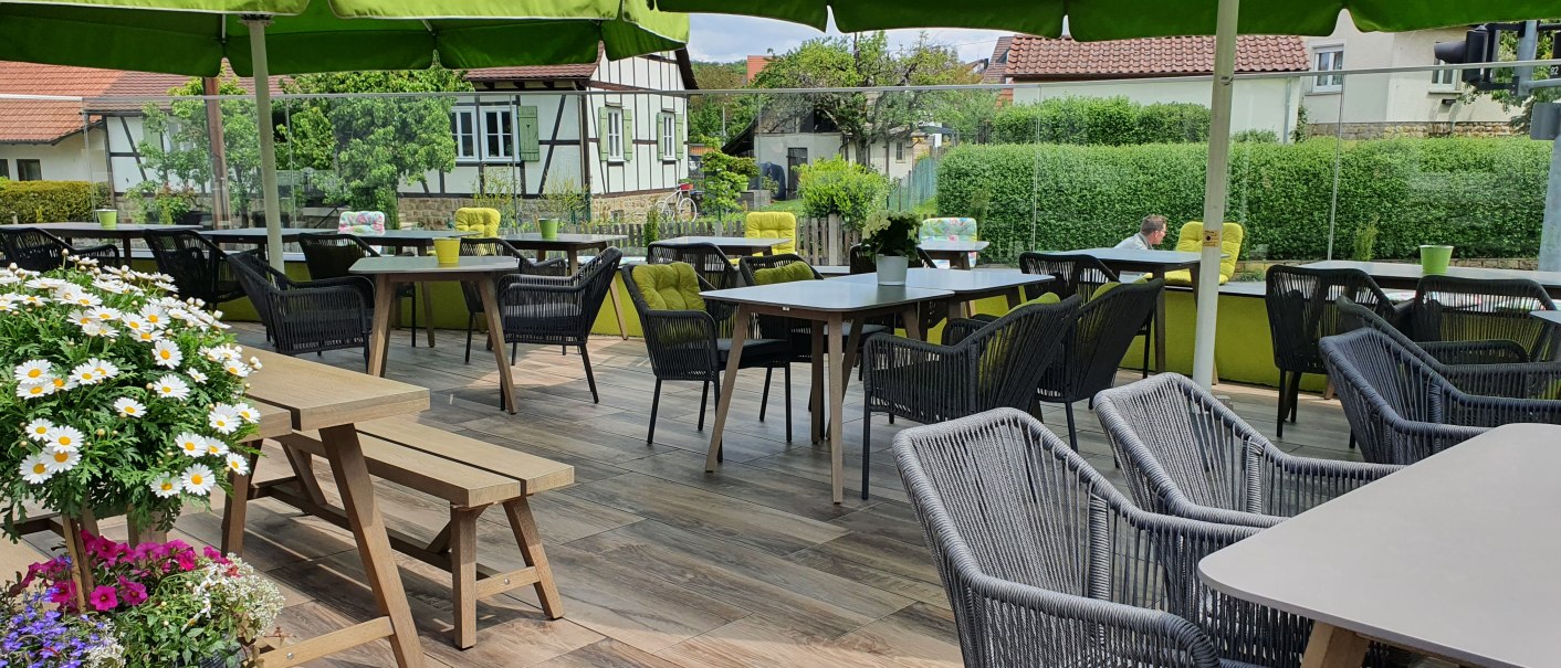 Gem&uuml;tliche Terrasse eines Landgasthofs mit gr&uuml;nen Sonnenschirmen, Holztischen und St&uuml;hlen. Blumen schm&uuml;cken den Bereich, im Hintergrund Fachwerkh&auml;user., &copy; Landgasthof Linde