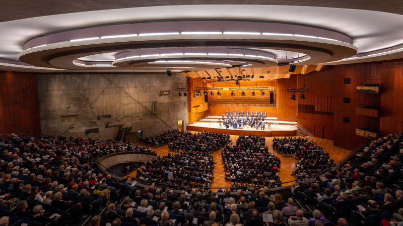 Ein großer Konzertsaal mit modernem Design, gefüllt mit Zuschauern. Auf der Bühne spielt ein Orchester. Die Architektur ist beeindruckend., © Holger Schneider Ein großer Konzertsaal mit modernem Design, gefüllt mit Zuschauern. Auf der Bühne spielt ein Orchester. Die Architektur ist beeindruckend., © Holger Schneider