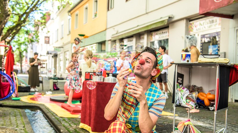 Ein Clown kniet in der Fußgängerzone und macht Seifenblasen. Im Hintergrund sind Kinder zu sehen., © Marcus Gauß Ein Clown kniet in der Fußgängerzone und macht Seifenblasen. Im Hintergrund sind Kinder zu sehen., © Marcus Gauß