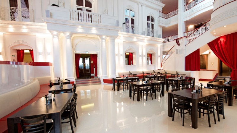 Elegantes Restaurant mit hohen weißen Wänden, roten Vorhängen und dunklen Holztischen. Die Architektur ist klassisch mit Säulen und Balkonen., © Dormero Hotel Stuttgart