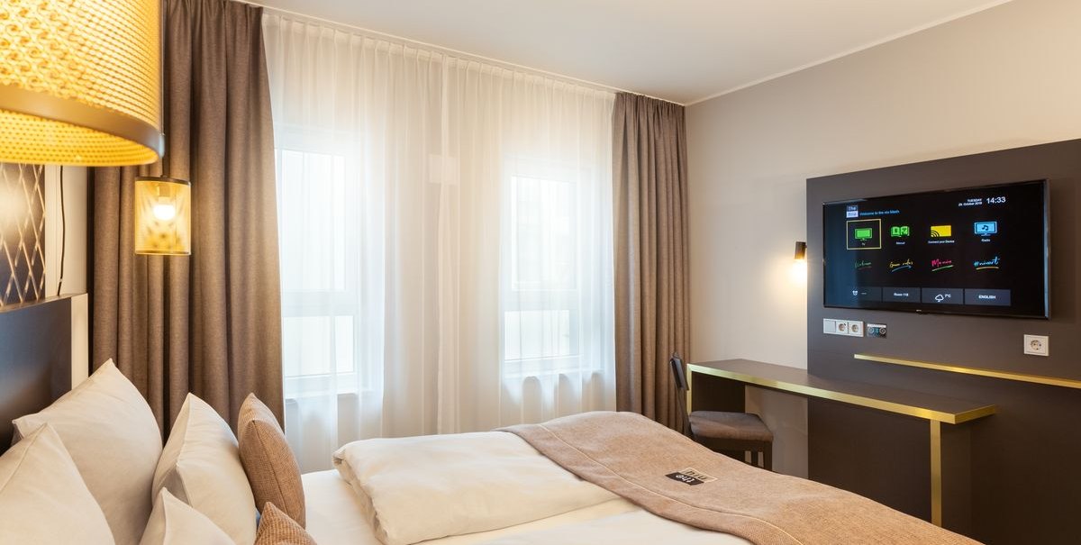 Modernes Hotelzimmer mit Doppelbett, TV an der Wand und kleinem Schreibtisch. Große Fenster mit Vorhängen lassen Tageslicht herein., © Novum Hospitality Modernes Hotelzimmer mit Doppelbett, TV an der Wand und kleinem Schreibtisch. Große Fenster mit Vorhängen lassen Tageslicht herein., © Novum Hospitality
