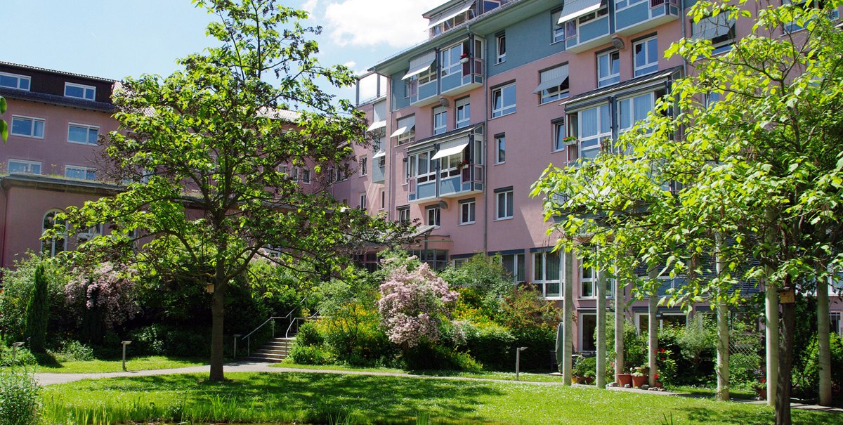 Mehrstöckiges Wohngebäude mit rosa und blauen Fassaden, umgeben von einem grünen Garten mit Bäumen und einem kleinen Teich., © Ev. Diakonissenanstalt Stuttgart Mehrstöckiges Wohngebäude mit rosa und blauen Fassaden, umgeben von einem grünen Garten mit Bäumen und einem kleinen Teich., © Ev. Diakonissenanstalt Stuttgart