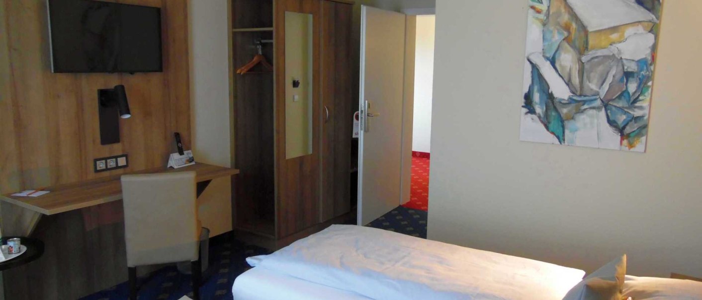 Ein Hotelzimmer mit einem Bett, Schreibtisch, Fernseher und einem abstrakten Kunstwerk an der Wand. Der Raum wirkt modern und aufgeräumt., © TOMAS Ein Hotelzimmer mit einem Bett, Schreibtisch, Fernseher und einem abstrakten Kunstwerk an der Wand. Der Raum wirkt modern und aufgeräumt., © TOMAS