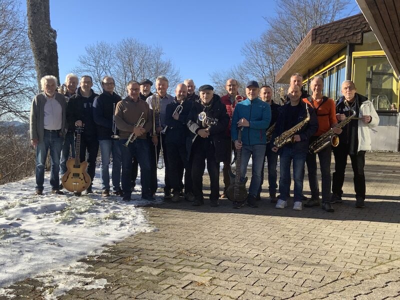 Die Herrenberg Bigband posiert mit Instrumenten im Freien vor einem Geb&auml;ude. Der Boden ist teils schneebedeckt, die Sonne scheint und B&auml;ume sind im Hintergrund sichtbar., &copy; Interessengemeinschaft Kultur Sindelfingen / B&ouml;blingen e.V.