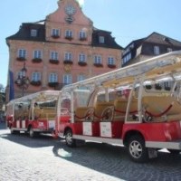 Ein roter Touristenzug steht auf einem gepflasterten Platz vor einem historischen Gebäude mit Uhr und Blumenkästen., © Für die Bildrechte: Amt für Medien und Kommunikation Schwäbisch Gmünd