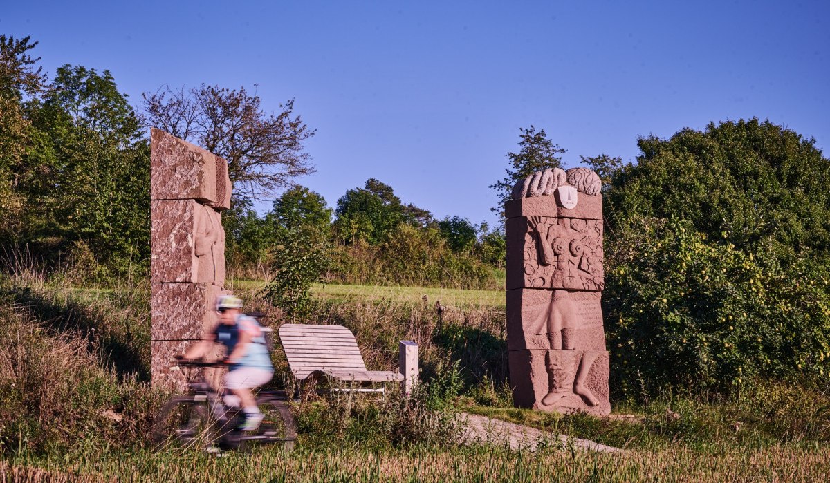 Ein Radfahrer passiert zwei große Steinskulpturen in einer grünen Landschaft unter klarem Himmel., © Natur.Nah. Schönbuch & Heckengäu Ein Radfahrer passiert zwei große Steinskulpturen in einer grünen Landschaft unter klarem Himmel., © Natur.Nah. Schönbuch & Heckengäu