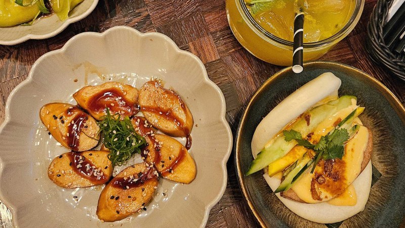 Ein Teller mit veganen Teigtaschen in So&szlig;e, ein Bao Bun mit Gem&uuml;se und ein Getr&auml;nk mit Eisw&uuml;rfeln auf einem Holztisch., &copy; SMG
