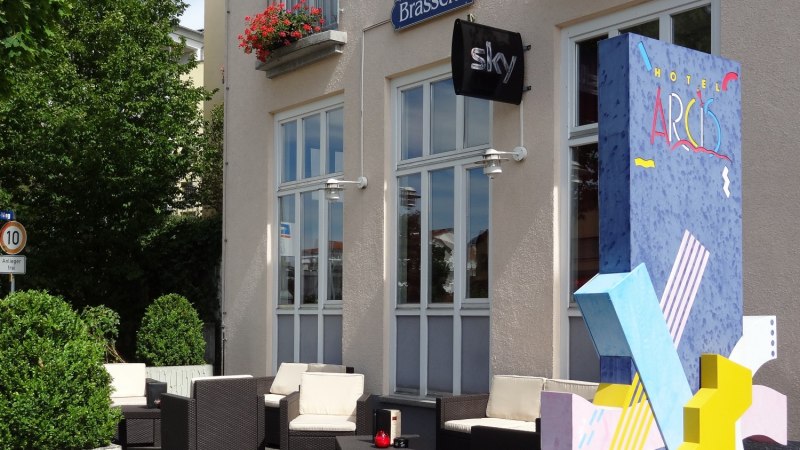 Terrasse des Hotels Arcis mit Sitzmöbeln und bunter Dekoration. Schilder für Brasserie und Sky sind sichtbar. Blumen schmücken die Fenster., © Hotel Arcis Terrasse des Hotels Arcis mit Sitzmöbeln und bunter Dekoration. Schilder für Brasserie und Sky sind sichtbar. Blumen schmücken die Fenster., © Hotel Arcis