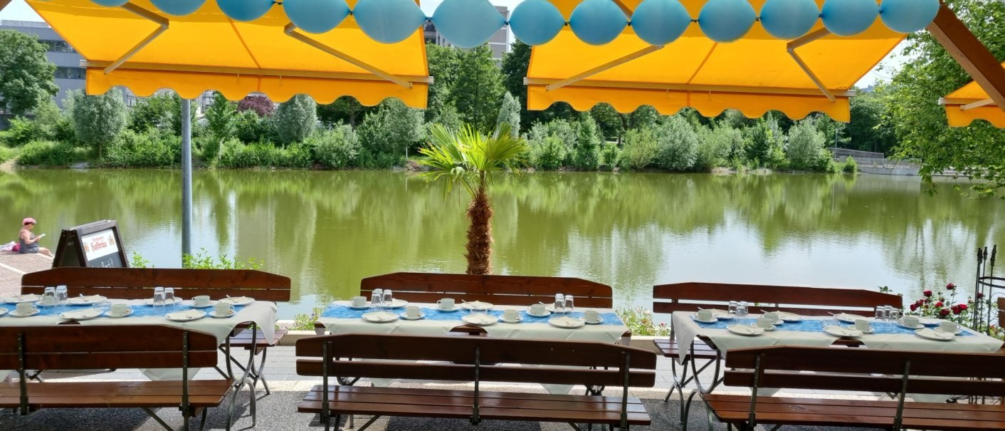 Gedeckte Tische unter gelben Markisen, dekoriert mit blauen Luftballons, mit Blick auf einen See und grüne Bäume im Hintergrund., © Gaststätte Goldbachsee Gedeckte Tische unter gelben Markisen, dekoriert mit blauen Luftballons, mit Blick auf einen See und grüne Bäume im Hintergrund., © Gaststätte Goldbachsee