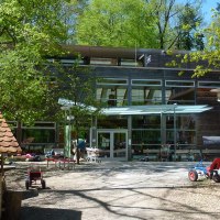 Innenhof des Haus des Waldes mit Spielzeugfahrzeugen, Bäumen und einem kleinen Holzhaus. Moderne Glasfassade im Hintergrund., © Haus des Waldes