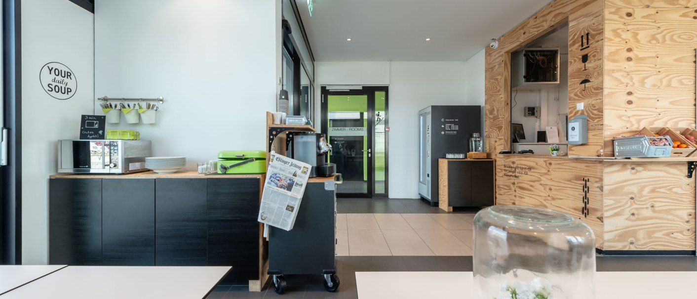 Moderne Hotel-Lounge mit Holzw&auml;nden, Snackbereich und Tisch mit Glasglocke. Zeitung und Kaffeemaschine sichtbar. Helle, einladende Atmosph&auml;re., &copy; a2 HOTELS