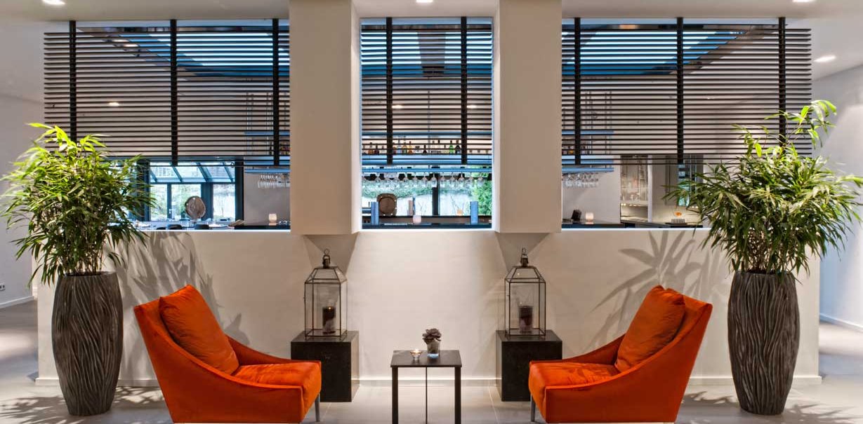 Moderne Lobby mit zwei orangefarbenen Sesseln, großen Pflanzen in Vasen und dekorativen Laternen auf schwarzen Tischen., © Wyndham Stuttgart Airport Messe
