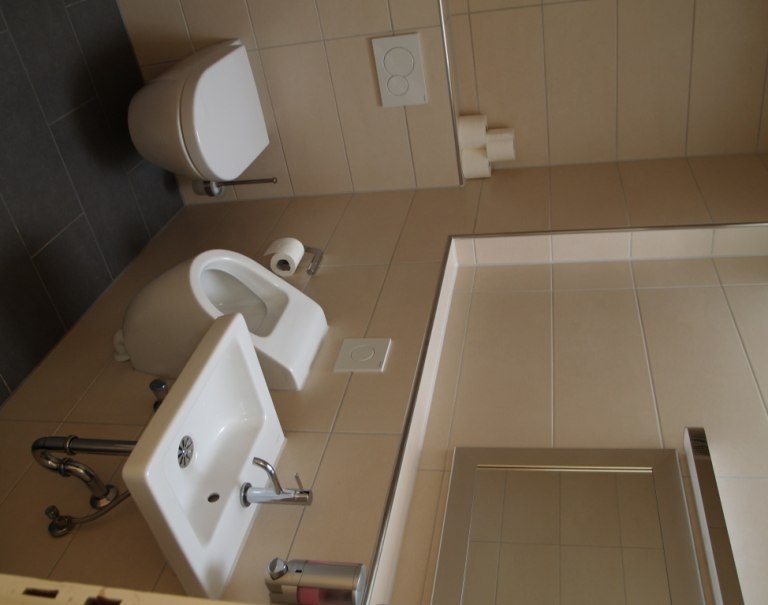 Ein modernes Badezimmer mit Toilette, Bidet und Waschbecken, alles in Weiß gehalten. Die Wände sind mit beigen Fliesen verkleidet., © TOMAS Ein modernes Badezimmer mit Toilette, Bidet und Waschbecken, alles in Weiß gehalten. Die Wände sind mit beigen Fliesen verkleidet., © TOMAS