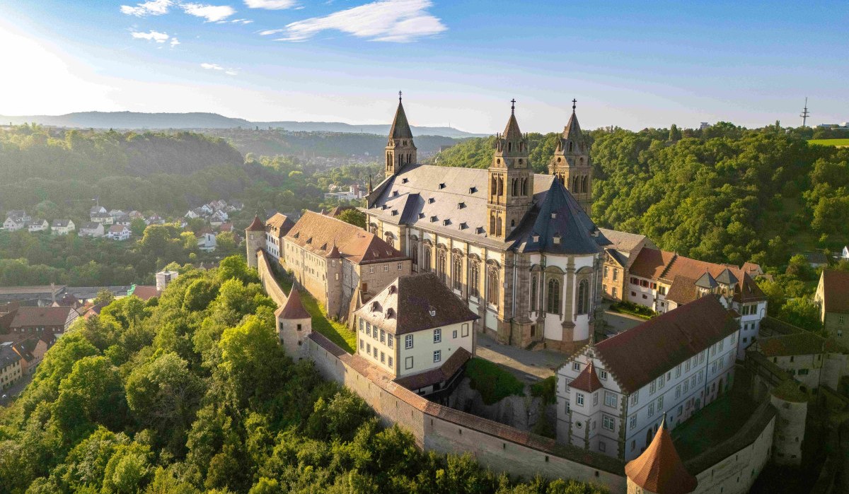 Luftaufnahme der Großcomburg in Schwäbisch Hall, umgeben von grünen Wäldern und Hügeln unter blauem Himmel., © Michael Kühneisen Luftaufnahme der Großcomburg in Schwäbisch Hall, umgeben von grünen Wäldern und Hügeln unter blauem Himmel., © Michael Kühneisen