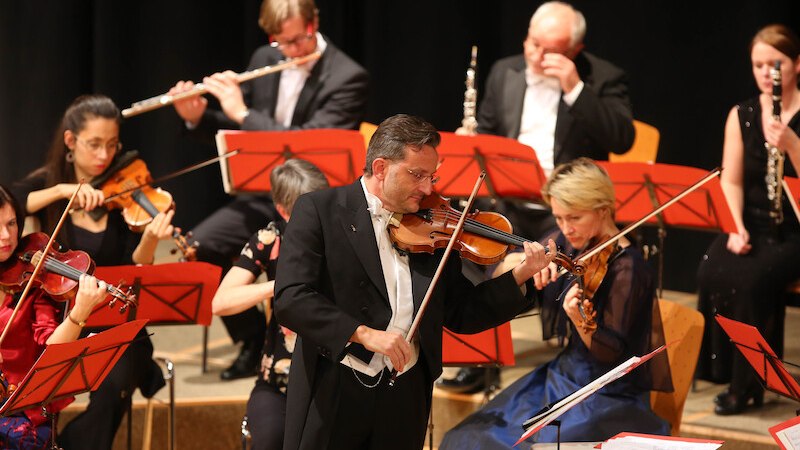 Ein Orchester spielt ein Konzert. Ein Geiger im Vordergrund, umgeben von Musikern mit Geigen und Blasinstrumenten. Alle tragen formelle Kleidung., &copy; Touristik Bad Wildbad GmbH
