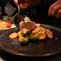 Ein Koch richtet ein Gericht mit Fleisch, Gemüse und Beilagen auf einem schwarzen Teller an. Im Hintergrund sind weitere Speisen zu sehen., © Bad Urach Tourismus