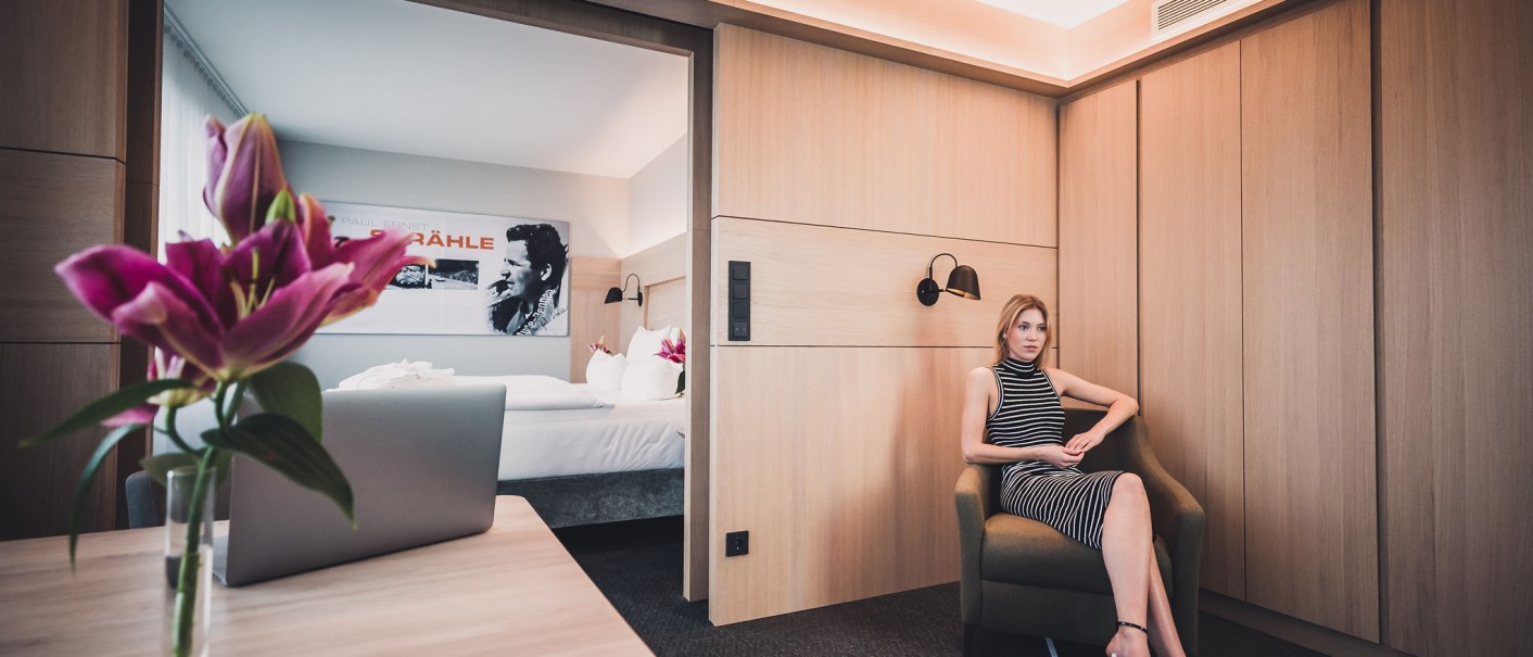 Eine Frau in einem gestreiften Kleid sitzt in einem modernen Hotelzimmer. Im Hintergrund sind ein Bett, Blumen und ein Bild an der Wand sichtbar., © Abacco Hotels GmbH Eine Frau in einem gestreiften Kleid sitzt in einem modernen Hotelzimmer. Im Hintergrund sind ein Bett, Blumen und ein Bild an der Wand sichtbar., © Abacco Hotels GmbH