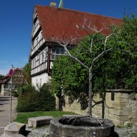 Brunnen mit Metallstruktur vor Fachwerkhaus und blauem Himmel. Historische Architektur und grüne Vegetation im Hintergrund., © RadL Leonberg