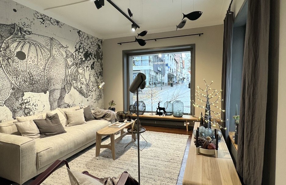 Modernes Wohnzimmer mit beigem Sofa, Kissen, Wandkunst, hängenden Fischen und dekorativen Elementen auf Tisch und Fensterbank., © schöner laden