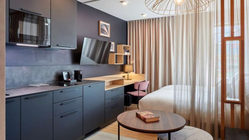 Modernes Studio mit dunkler Küche, Schreibtisch, Bett und stilvoller Beleuchtung. Vorhänge trennen den Schlafbereich ab., © Adagio Modernes Studio mit dunkler Küche, Schreibtisch, Bett und stilvoller Beleuchtung. Vorhänge trennen den Schlafbereich ab., © Adagio