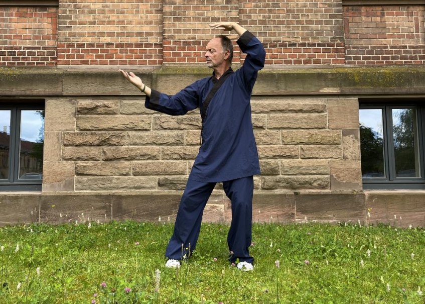 Eine Person in blauer Kleidung f&uuml;hrt eine Taiji Qigong-&Uuml;bung vor einer Backsteinmauer aus. Sie steht auf einer gr&uuml;nen Wiese., &copy; &copy; Felina Konkart