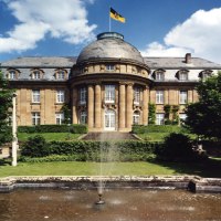 Die Villa Reitzenstein mit imposanter Fassade, einem Brunnen im Vordergrund und einer Flagge auf dem Dach, umgeben von Bäumen., © Staatsministerium Baden-Württemberg Die Villa Reitzenstein mit imposanter Fassade, einem Brunnen im Vordergrund und einer Flagge auf dem Dach, umgeben von Bäumen., © Staatsministerium Baden-Württemberg