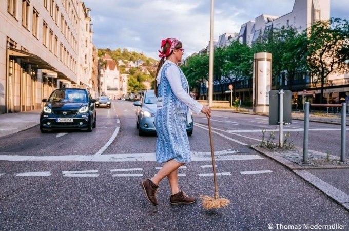 Eine Frau in traditioneller Kleidung mit Kopftuch und Besen &uuml;berquert eine Stra&szlig;e in einer Stadt. Autos sind im Hintergrund zu sehen., &copy; Stuttgart Marketing GmbH