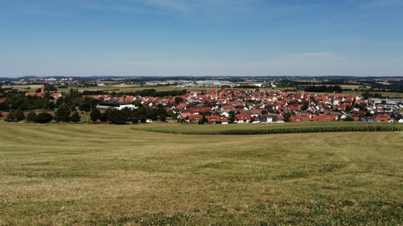 Grüne Wiesen führen zu einer Siedlung mit roten Dächern, umgeben von Feldern und Wäldern unter blauem Himmel., © Foto: Cornelia Steinbach Grüne Wiesen führen zu einer Siedlung mit roten Dächern, umgeben von Feldern und Wäldern unter blauem Himmel., © Foto: Cornelia Steinbach