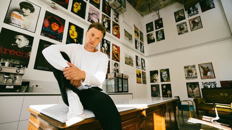 Eine Person sitzt l&auml;ssig auf einem Tisch in einem Raum voller bunter Filmplakate. Die Atmosph&auml;re wirkt kreativ und k&uuml;nstlerisch., &copy; Kulturstiftung Hohenlohe