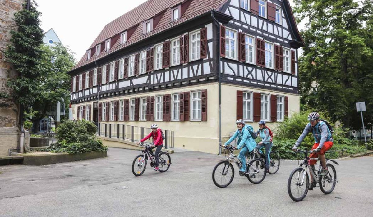 Vier Radfahrer fahren an einem historischen Fachwerkhaus in Kirchheim unter Teck vorbei. Das Gebäude hat weiße Wände und braune Fensterläden., © Schwäbische Alb Tourismusverband e.V.