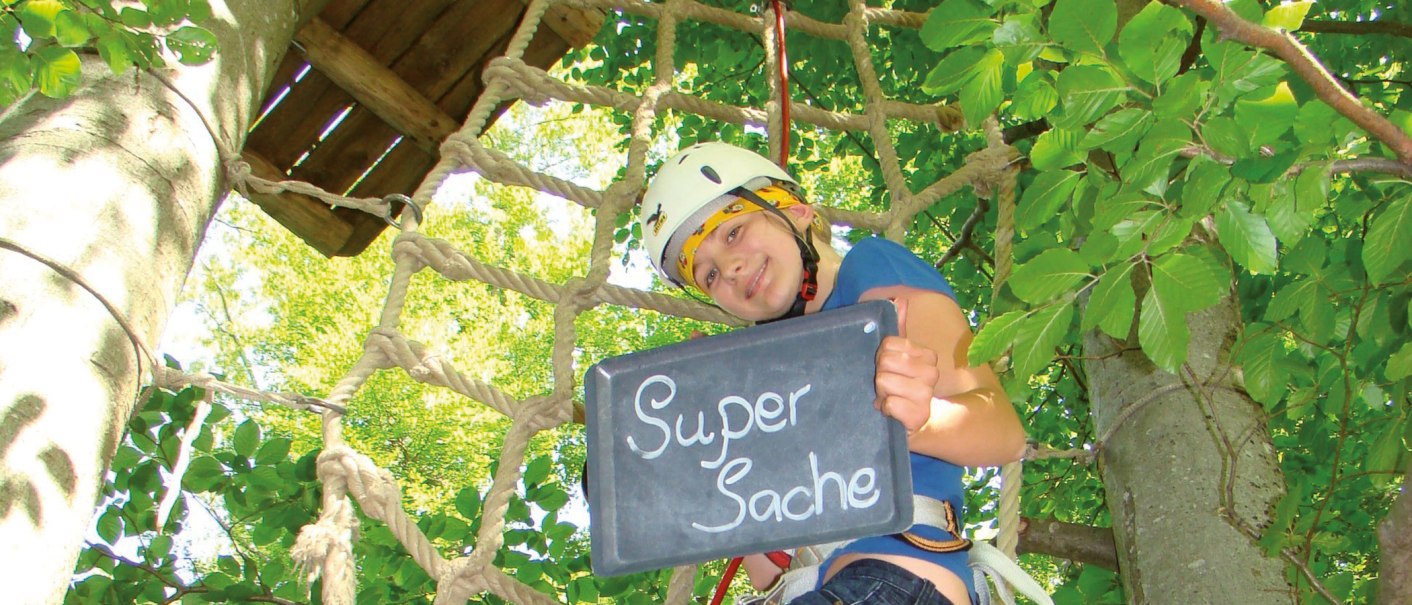 Person im Kletterpark hält eine Tafel mit der Aufschrift 'Super Sache' und trägt einen Helm. Im Hintergrund sind Bäume und ein Kletternetz zu sehen., © Stuttgart-Marketing GmbH