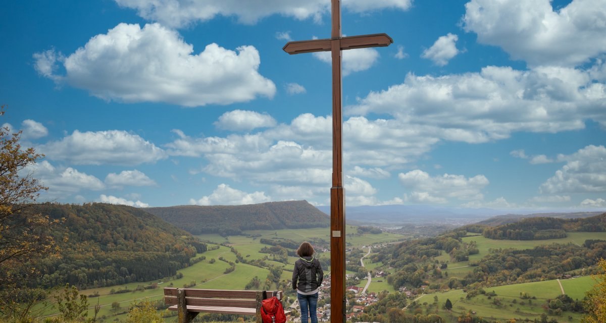 Person am Aussichtspunkt Kreuz Galgenberg, neben einem großen Holzkreuz, mit Blick auf eine weite grüne Landschaft und einen bewölkten blauen Himmel., © Foto: Mario Klaiber Person am Aussichtspunkt Kreuz Galgenberg, neben einem großen Holzkreuz, mit Blick auf eine weite grüne Landschaft und einen bewölkten blauen Himmel., © Foto: Mario Klaiber