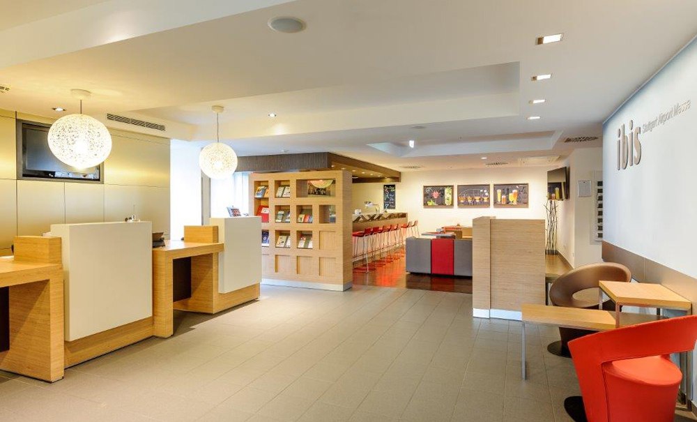 Moderne Hotelrezeption mit Holztheken, Sitzbereich und dekorativen Lampen. Im Hintergrund ein Bücherregal und eine Bar mit roten Hockern., © Ibis Stuttgart Airport Messe Moderne Hotelrezeption mit Holztheken, Sitzbereich und dekorativen Lampen. Im Hintergrund ein Bücherregal und eine Bar mit roten Hockern., © Ibis Stuttgart Airport Messe