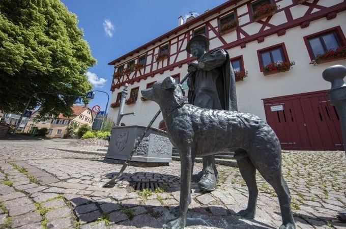 Bronzestatue eines Hirten mit Hund vor einem Fachwerkhaus in Schafhausen. Der gepflasterte Platz und blauer Himmel erg&auml;nzen die Szene., &copy; Stadtverwaltung Weil der Stadt
