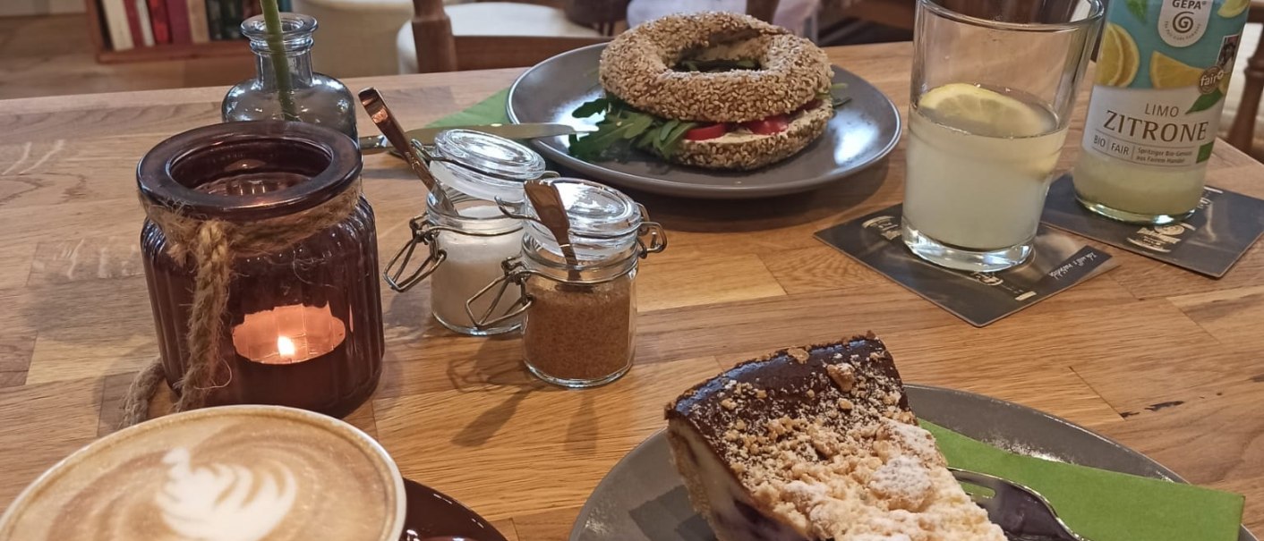 Gem&uuml;tliches Caf&eacute; mit Cappuccino, Kuchen und Bagel auf einem Holztisch. Im Hintergrund B&uuml;cherregale und bequeme Sessel., &copy; cKaffeehausMaLe