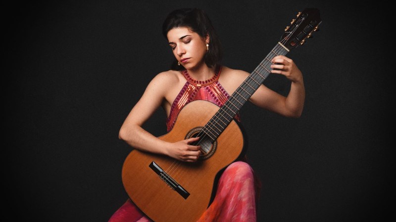 Eine Frau in einem bunten Kleid spielt Gitarre vor einem dunklen Hintergrund. Sie wirkt konzentriert und vertieft in die Musik.