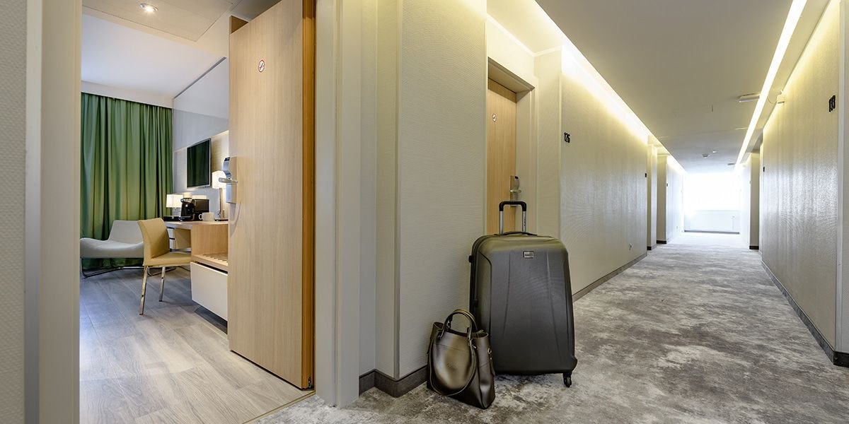 Ein Hotelkorridor mit Teppichboden. Ein Zimmer ist offen, innen sind grüne Vorhänge sichtbar. Ein Koffer und eine Tasche stehen vor der Tür., © Mercure Bristol Stuttgart Sindelfingen