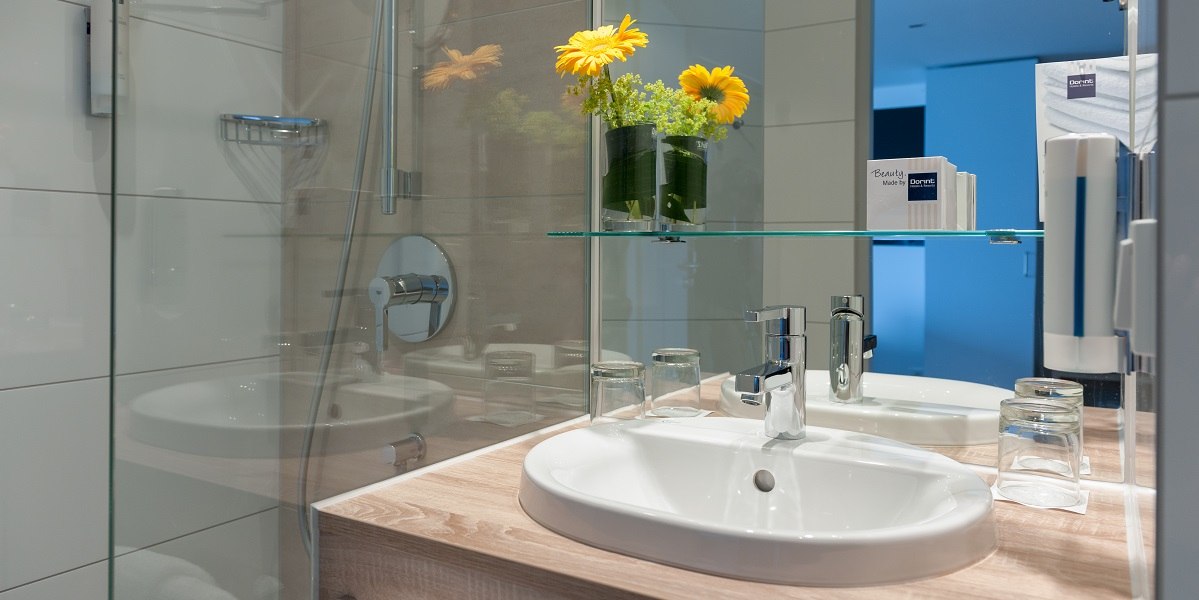Modernes Badezimmer mit Waschbecken, Spiegel, Glasregal und gelben Blumen. Glasdusche und Pflegeprodukte sind ebenfalls sichtbar., © Essential by Dorint