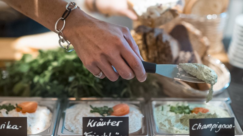 Eine Hand mit Armband nimmt Champignoncreme aus einer Schale. Daneben stehen Schalen mit Kr&auml;uterfrischk&auml;se und Paprikacreme, im Hintergrund Brot., &copy; Caf&eacute; Justus, Stuttgart