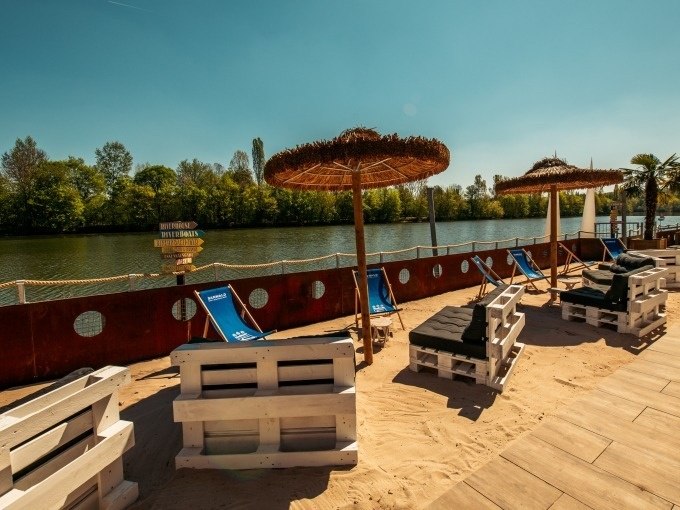 Strandbereich mit Liegestühlen, Sonnenschirmen und Palettenmöbeln am Flussufer. Im Hintergrund sind Bäume und ein klarer Himmel zu sehen., © Cool-Tours StattReisen Strandbereich mit Liegestühlen, Sonnenschirmen und Palettenmöbeln am Flussufer. Im Hintergrund sind Bäume und ein klarer Himmel zu sehen., © Cool-Tours StattReisen
