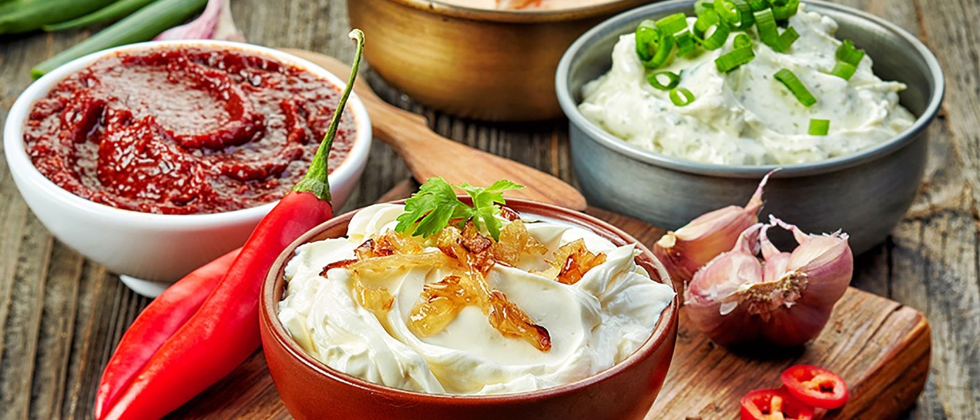 Verschiedene Dips in Schalen, umgeben von roten Chilischoten und Knoblauch auf einem Holztisch arrangiert., © MASEVEN Stuttgart SI-Centrum Verschiedene Dips in Schalen, umgeben von roten Chilischoten und Knoblauch auf einem Holztisch arrangiert., © MASEVEN Stuttgart SI-Centrum