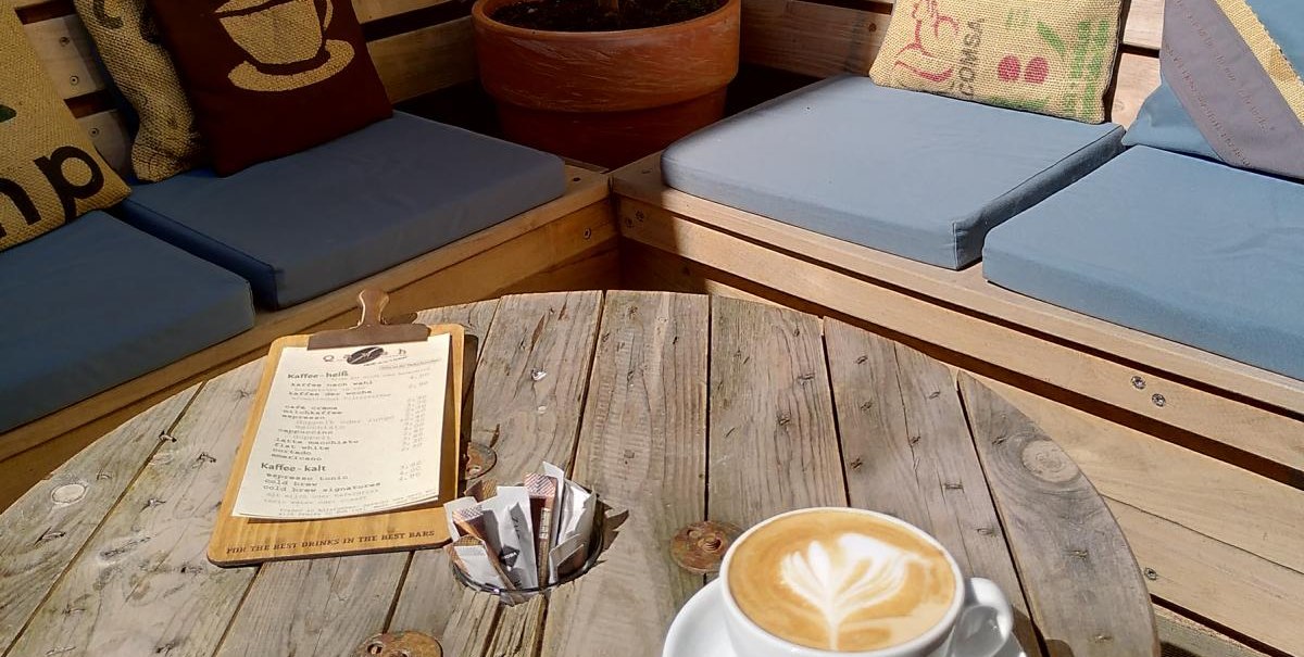 Gemütlicher Außenbereich mit Holzmöbeln, Kissen und Pflanzen. Auf dem Tisch steht eine Tasse Cappuccino und eine Speisekarte., © Matthias Gohl Gemütlicher Außenbereich mit Holzmöbeln, Kissen und Pflanzen. Auf dem Tisch steht eine Tasse Cappuccino und eine Speisekarte., © Matthias Gohl