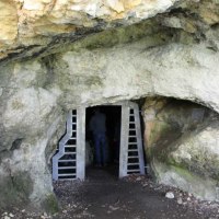 Höhleneingang mit Metallgitter, eine Person steht im Inneren. Die Höhle ist von Felsen umgeben.