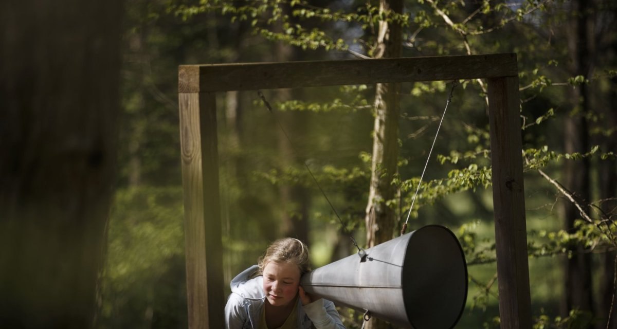 Ein Mädchen lauscht einem großen Metalltrichter, der an einem Holzrahmen im Wald befestigt ist. Die Umgebung ist grün und bewaldet., © Natur.Nah. Schönbuch & Heckengäu Ein Mädchen lauscht einem großen Metalltrichter, der an einem Holzrahmen im Wald befestigt ist. Die Umgebung ist grün und bewaldet., © Natur.Nah. Schönbuch & Heckengäu