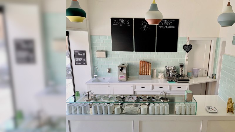 Eisdiele mit Theke, Eismaschine, Kaffeebereiter und Eiswaffeln. Pastellfarbene Fliesen und Lampen. Schwarze Tafeln mit Eisangeboten., © Zur Schleckerei Eisdiele mit Theke, Eismaschine, Kaffeebereiter und Eiswaffeln. Pastellfarbene Fliesen und Lampen. Schwarze Tafeln mit Eisangeboten., © Zur Schleckerei