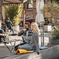 Menschen genießen Kaffee vor einem Café in der Sonne. Ein Paar sitzt auf einer Bank, andere an Tischen. Pflanzen und Sonnenschirme schmücken die Szene., © SMG, Sarah Schmid