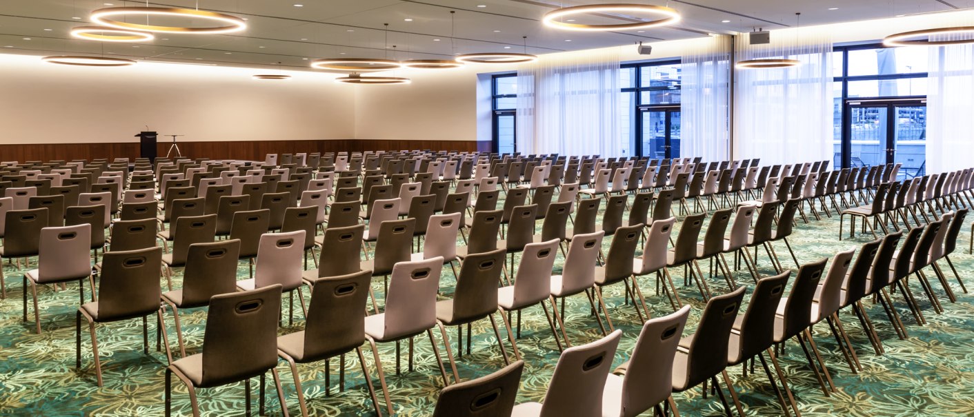 Ein leerer Ballsaal mit vielen Stuhlreihen, modernen Ringlichtern an der Decke und einem Rednerpult vorne. Der Boden hat ein grünes Muster., © Mövenpick Hotel Stuttgart Messe & Congress Ein leerer Ballsaal mit vielen Stuhlreihen, modernen Ringlichtern an der Decke und einem Rednerpult vorne. Der Boden hat ein grünes Muster., © Mövenpick Hotel Stuttgart Messe & Congress