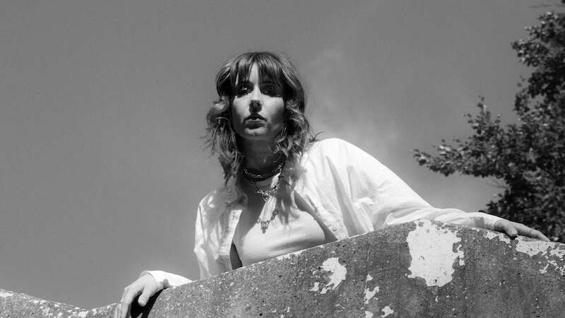 Schwarz-weiß Foto einer Person mit welligem Haar, die über eine Betonmauer schaut. Der Himmel ist klar, und Bäume sind im Hintergrund sichtbar., © Kulturhaus Schwanen