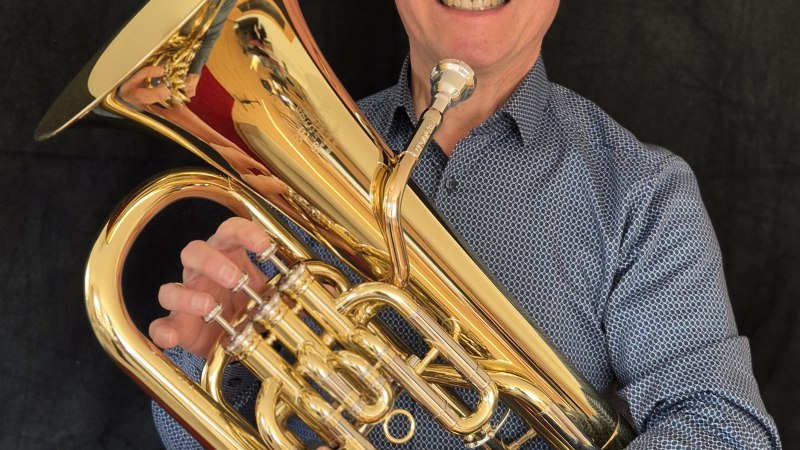 Ein Mann mit Brille und blauem Hemd h&auml;lt ein gl&auml;nzendes Euphonium vor einem dunklen Hintergrund und l&auml;chelt in die Kamera., &copy; Gert K&uuml;rner